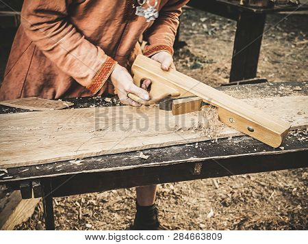 Carpenter In Medieval Cotton Clothes Working With A Wood By Plane. Man Manually Pours Sawdust From T