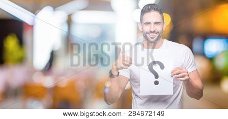 Handsome young man holding paper with question mark over isolated background happy with big smile doing ok sign, thumb up with fingers, excellent sign