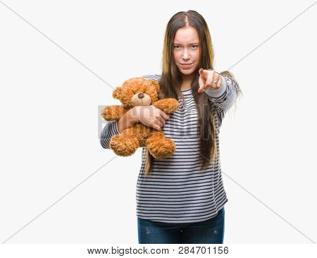 Young caucasian woman holding teddy bear over isolated background pointing with finger to the camera and to you, hand sign, positive and confident gesture from the front