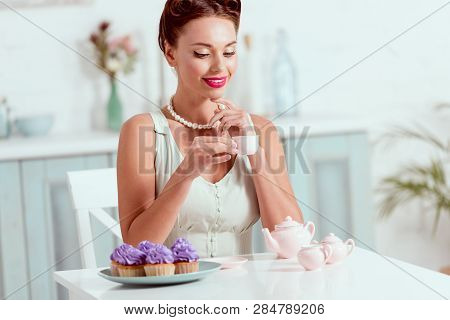 Pritty Pin Up Girl Holding Cup Of Coffee While Sitting At Table With Plate Of Cupcakes