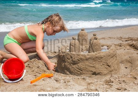 Little girl wearing green swimsuitbuilding a sand cactle on a sandy beach in Crete, Greece. Family summer vacation concept