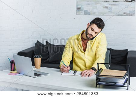 Concentarted Bearded Businessman Talking On Smartphone And Writing In Notebook