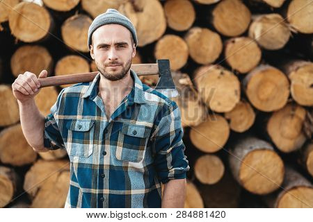 Strong Bearded Lumberman Wearing Plaid Shirt Holds Axe On His Shoulder On Background Of Sawmill