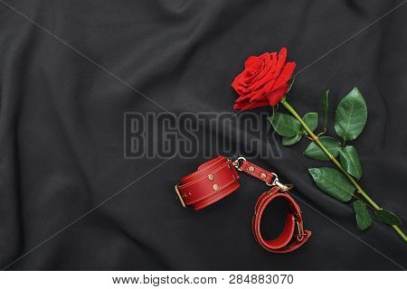 Bdsm Fantasy Play. Top View Of Red Leather Handcuffs And Rose Against Of Black Silk Fabric