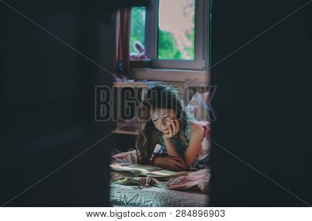 Thoughtful Kid Girl Reading Book Alone In Her Room In The Veening Or Early Morning
