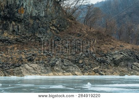 Wall Of Volcanic Rock On Shore Of Frozen River With Background Of Dense Trees On Winter Day.