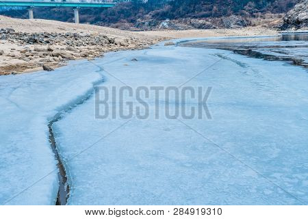 Long Crack In Ice Sheet On Frozen River With Sandy Rocky Shore With Bridge In Background On Winter M