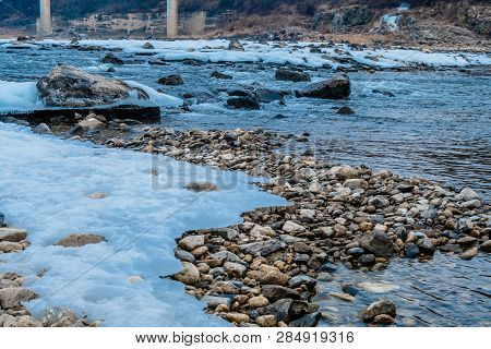 Rapidly Flowing River With Rocky Riverbed And Rocky Shoreline Covered With Sheets Of Ice Snow.