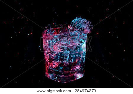 Splash And Droplets Of Liquid In A Rocks Glass Under Colorful Lighting Isolated On A Black Backgroun