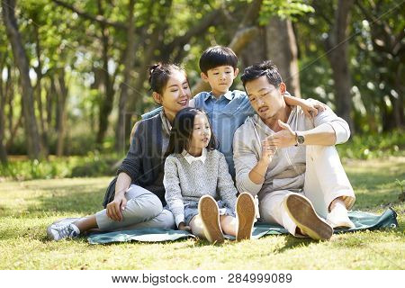 Asian Family With Two Children Having Fun Sitting On Grass Talking Chatting Outdoors In Park