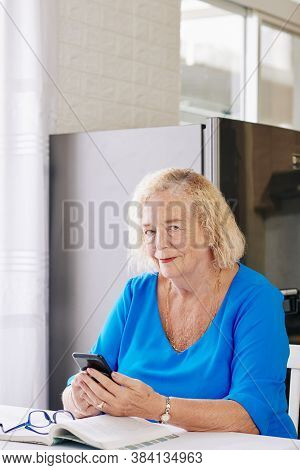 Portrait Of Smiling Elderly Woman Sitting At Desk In Kitchen And Answering Text Messages From Her Fr