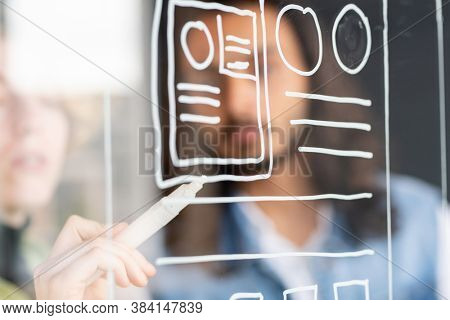 Hand of young female developer of mobile application holding white highlighter by transparent noteboard while drawing sketch