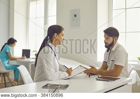 Medical Clinic. Smiling Woman Doctor Sitting And Listening To Talking Man Patient During Consultatio