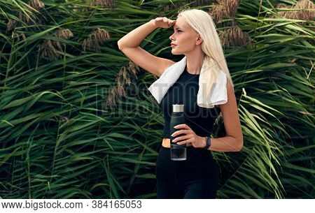 Outdoor Horizontal Shot Of Beautiful Blonde Female Runner Standing Against Nature Background Holding
