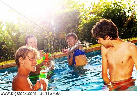 Four Happy Laughing Friends Play With Water-gun Squirt Pistol On Sunny Day In Swimming Pool