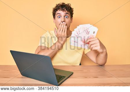 Young caucasian man with curly hair sitting on the table working with laptop and holding israel shekels banknotes covering mouth with hand, shocked and afraid for mistake. surprised expression 