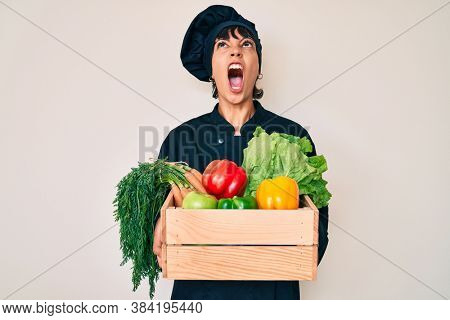 Beautiful brunettte woman chef holding fresh veggetables angry and mad screaming frustrated and furious, shouting with anger looking up. 