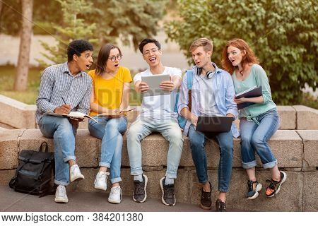 Asian Student Guy Showing His Project On Digital Tablet To Fellow-students Sitting Outdoors. College
