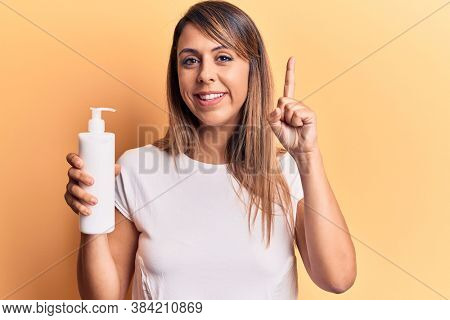 Young beautiful woman holding bottle of cream lotion smiling with an idea or question pointing finger with happy face, number one 