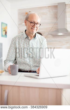 Old Man Smiling While Watching A Movie On The Laptop In The Kitcen. Daily Life Of Senior Man In Kitc
