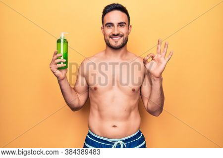 Handsome man with beard on vacation wearing swimwear holding bottle of aloe vera cream doing ok sign with fingers, smiling friendly gesturing excellent symbol