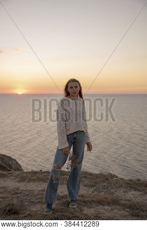 A Blonde Girl In Leaky Jeans Poses Against The Background Of The Sunset .