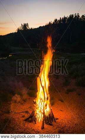 Midsummer Ligo Celebration. Huge Bonfire On The River Shore. Scenic Landscape Of Forest. Sunset Sky.