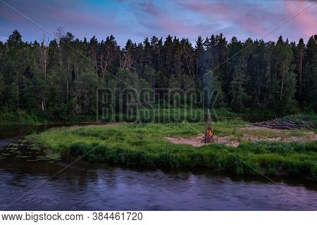 Midsummer Ligo Celebration. Huge Fire On The River Shore. Scenic Landscape Of Forest At Twilight.