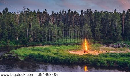 Traditional Midsummer Ligo Celebration. Huge Fire On The River Shore. Scenic Landscape Of Forest At 