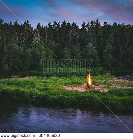 Midsummer Ligo Celebration. Huge Fire On The River Shore. Scenic Landscape Of Forest At Twilight.