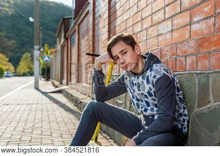 A Teenage Boy Sits On The Sidewalk Next To A Scooter And Poses Leaning On His Arm. Brick Wall And St
