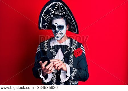 Young man wearing mexican day of the dead costume using smartphone looking positive and happy standing and smiling with a confident smile showing teeth 