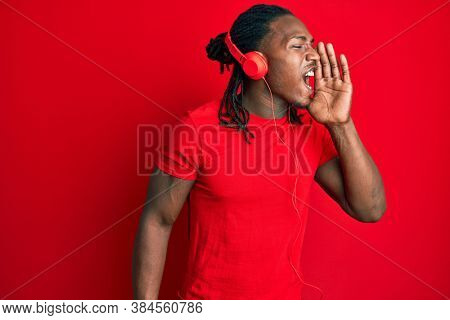 African american man with braids listening to music using headphones shouting and screaming loud to side with hand on mouth. communication concept. 