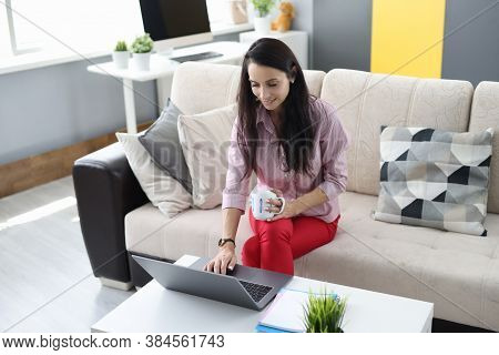 Woman Is Sitting On The Couch Holding Cup In Crayfish And With Her Other Hand Typing On Laptop Keybo