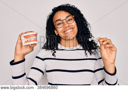 Young beautiful african american woman with curly hair smiling happy and confident. Standing with smile on face holding plastic denture teeth and invisible aligner over isolated white background