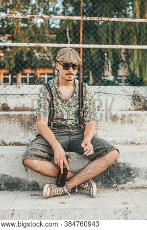 Young Hipster Man In Retro Clothing Sits On The Stairs With Beer In His Hands