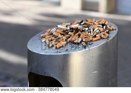 Many Old Cigarette Stubs Spilling Over From Ashtray Of Overflowing Public Trash Bin
