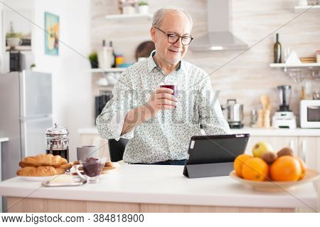 Elderly Man Using Tablet Pc In Kitchen During Breakfast Wearing Glasses. Elderly Person With Tablet 