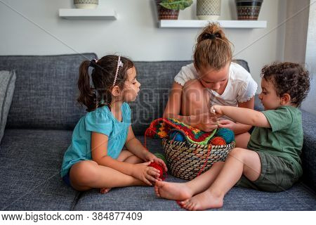 Children Learning To Knit, Sitting On The Couch In The Home. Two Girls And A Funny Curly Boy Knits C