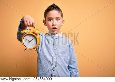 Young little boy kid holding classic bell alarm clock over isolated yellow background scared in shock with a surprise face, afraid and excited with fear expression