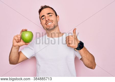 Young handsome man with blue eyes eating healthy green apple over isolated pink background happy with big smile doing ok sign, thumb up with fingers, excellent sign