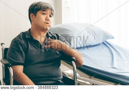 Asian Middle Aged Man  Sitting On A Wheelchair Close To The Patient Bed, His Hands Are Kinking Due T
