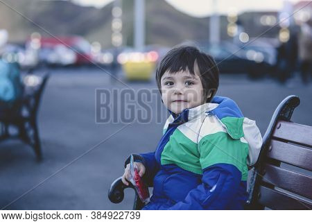 Potrait Of Happy Kid Siting On Wooden Bench With Burry Car Park Background, Boy Looking At Cameara W