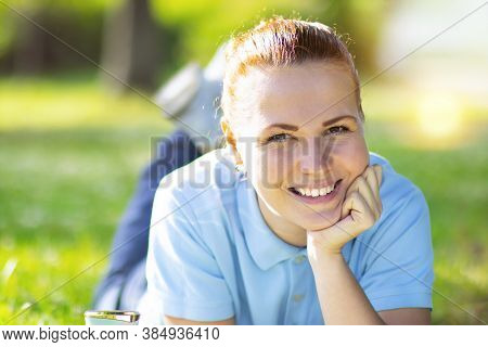 Happy Woman, Lies And Rests On A Green Lawn. The Female Is Smiling Behind The Grass In The Park. Out