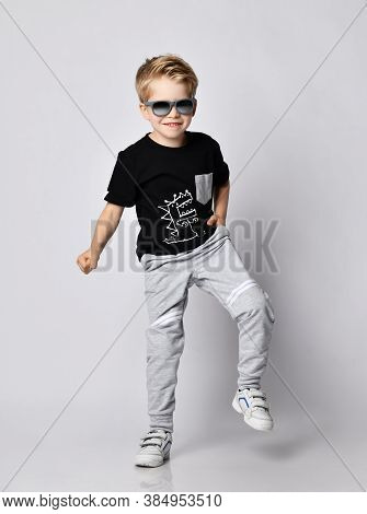 Frolic Blond Kid Boy In Sunglasses, Black T-shirt With Dinosaur Print And Gray Pants Stands With His