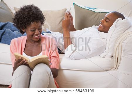 Happy couple relaxing together reading book and using smartphone at home in the living room