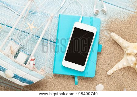 Smartphone and notepad on sea sand with starfish and toy boat. Top view with copy space