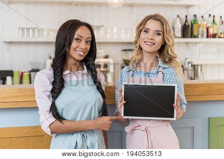 smiling beautiful multiethnic waitresses with blank board looking at camera while standing at cafe
