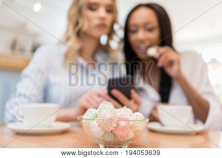 Young multiethnic women sitting at cafe and using smartphone together selective focus on cookies