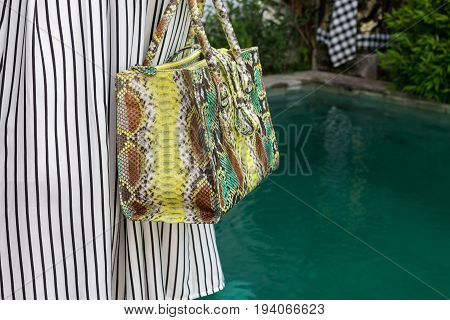 Fashionable woman holding luxury snakeskin python bag. Elegant outfit. Close up of purse in hands of stylish lady. Model posing near the swimming pool.